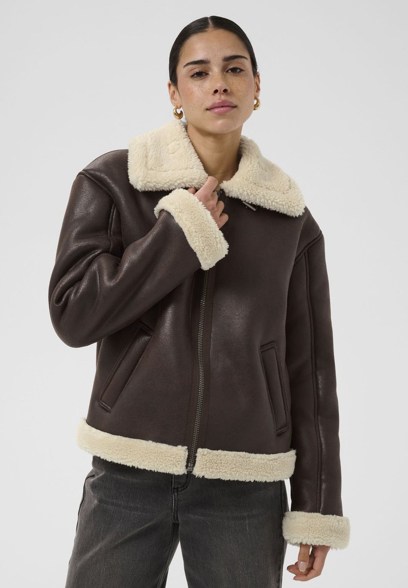 Femme portant une veste en cuir marron foncé avec un col, des poignets et un ourlet en shearling crème, debout devant un fond clair uni.