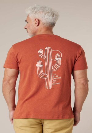 Homme d'âge moyen aux cheveux blancs portant un T-shirt couleur rouille avec un motif de cactus blanc et le texte « C'est mieux sous le soleil ! Deluxe » au dos.