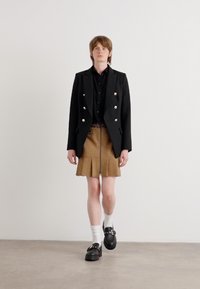 Schwarzes, doppelt geknöpftes Blazer mit silbernen Knöpfen, kombiniert mit einem braunen, plissierten Minirock. Schwarze Plateauschuhe und weiße Socken runden das Outfit ab.