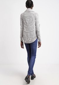 Grå stickad cardigan med franjkant, öppen ryggdesign med cirkulära mönster, åtsedd med mörkblå jeans och svarta skor.