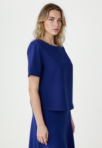 Blondynka o niebieskich oczach w królewskim niebieskim bluzce z krótkim rękawem i dopasowanej spódnicy, stojąca na jednolitym jasnym tle.