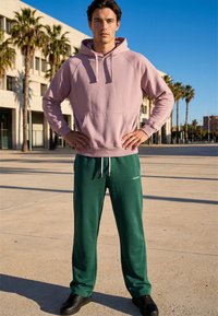 Un jeune homme se tient dehors, les mains sur les hanches, portant un sweat à capuche violet clair, un pantalon de survêtement vert foncé et des baskets noires sous un ciel bleu clair.