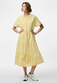 Vestido de algodón amarillo con un diseño de volantes, mangas cortas y un escote con cordón. Combinado con zapatillas blancas.