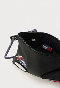 Musta kangaskassi vetoketjulla, jossa on monivärinen logoplaketti ja sininen kuvioitu olkahihna. Sisäpuolella pieni vetoketjutasku.