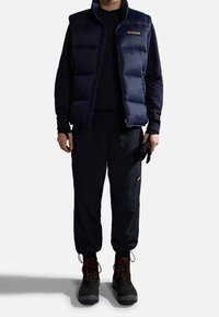 Gilet imbottito blu navy con zip frontale, abbinato a pantaloni scuri e stivali neri. Guanti tenuti nella mano sinistra. Abbigliamento outdoor semplice e funzionale.