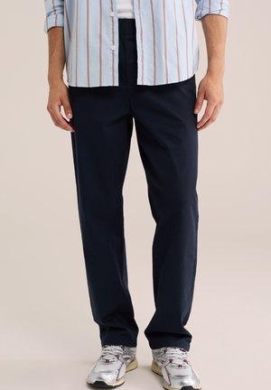 Homme portant un pantalon noir à taille élastique, des chaussures de course blanches et une chemise bleu clair avec des rayures rouges, debout devant un fond uni.