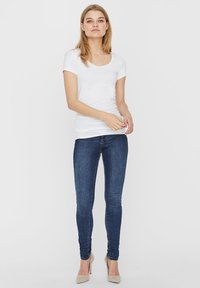 Witte scoopneck t-shirt gecombineerd met strakke blauwe jeans. Het model staat op beige hoge hakken en toont een eenvoudige, minimalistische stijl.