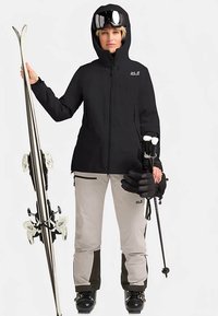 Veste de ski noire, pantalon de ski beige avec accents foncés, gants noirs, lunettes de ski sur la tête, tenant des skis et des bâtons, debout sur fond blanc.