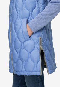 Veste matelassée bleu clair avec une finition texturée, dotée de poches latérales zippées et d'une coupe droite. Les manches sont en tricot bleu contrastant.