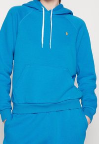 Ljusblå huvtröja gjord av mjukt tyg, med en främre ficka, vita snören och en liten orange logotyp på bröstet.