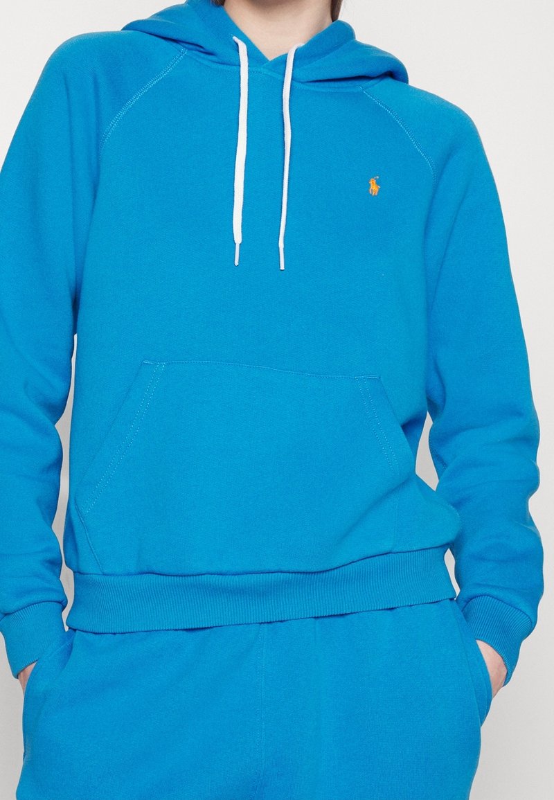 Ljusblå huvtröja gjord av mjukt tyg, med en främre ficka, vita snören och en liten orange logotyp på bröstet.