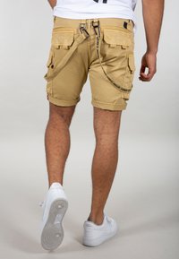 Khaki cargo šortky s viacerými vreckami, srolovanými manžetami a odnímateľným popruhom, kombinované s bielymi teniskami, na neutrálne pozadí.