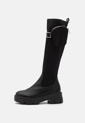 Musta polvipituiset saappaat, joissa on paksut kumipohjat, sileistä nahkaa oleva kärki, tekstiilipäällinen ja pieni vetoketjullinen tasku sivussa.