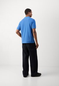 Blue cotton polo shirt with a classic collar, short sleeves, side slits, paired with loose-fitting black pants and footwear.