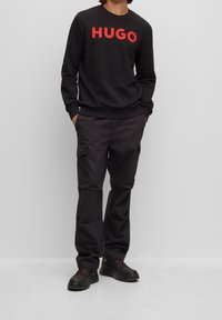 Sudadera negra con logo rojo "HUGO", combinada con pantalones cargo negros con bolsillos laterales. El calzado es negro con acentos rojos.