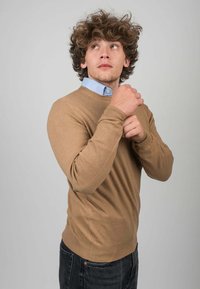 Bruine trui van zacht materiaal, met een ronde halslijn en lange mouwen, gelaagd over een lichtblauw overhemd met kraag, gecombineerd met donkere jeans.