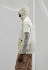 Lightweight, translucent cream rain jacket with a hood and elastic cuffs, paired with beige cargo pants featuring side pockets.