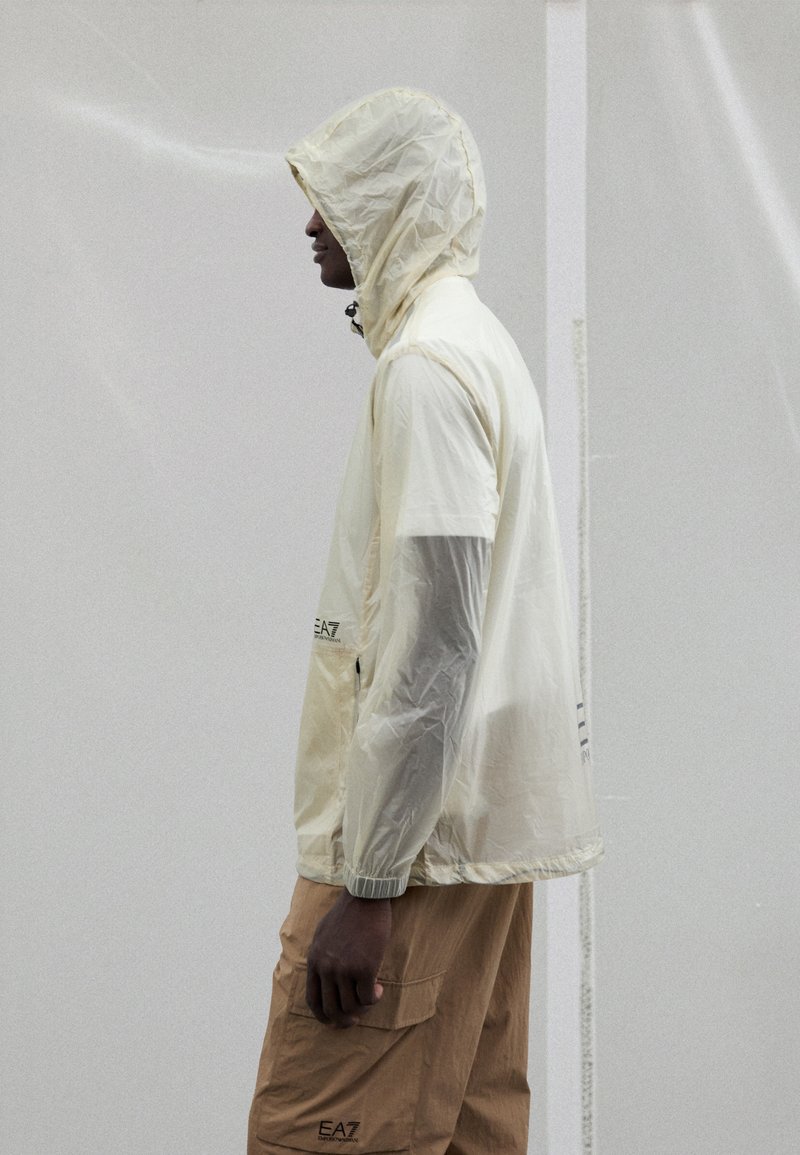 Lightweight, translucent cream rain jacket with a hood and elastic cuffs, paired with beige cargo pants featuring side pockets.
