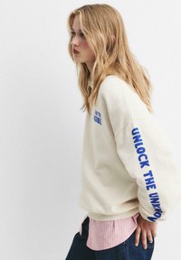 Blonde Frau in weißem Sweatshirt mit blauer Schrift "UNLOCK THE UNKNOWN" auf dem Ärmel, pink gestreiftem Hemd und dunkler Jeans, die nach links schaut.