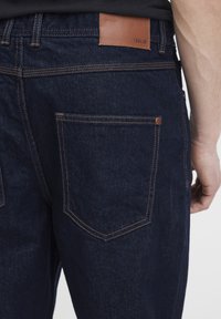 Temno modre kavbojke iz jeansa imajo strukturiran dizajn s kontrastnim šivanjem, usnjenim trakom na pasu in standardnimi zadnjimi žepi z zaobljenimi robovi.