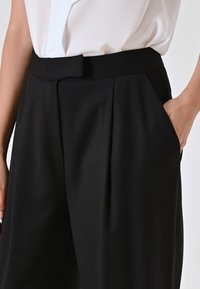 Zwarte op maat gemaakte broek met een vlakke tailleband, zijzakken, subtiele plooien en een gladde textuur, gecombineerd met een witte blouse.