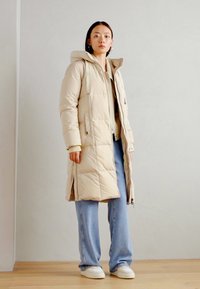 Beige lange pufferjas met een capuchon, rits aan de voorkant en zijzakken; gecombineerd met lichtblauwe jeans en witte sneakers. Glad materiaal.