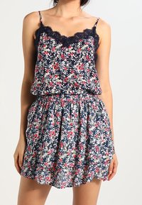 Kwiatowa sukienka z granatowym tłem, ozdobiona małymi czerwonymi i białymi kwiatkami. Na ramiączkach spaghetti, z koronkowym wykończeniem i marszczoną talią dla swobodnego kroju.