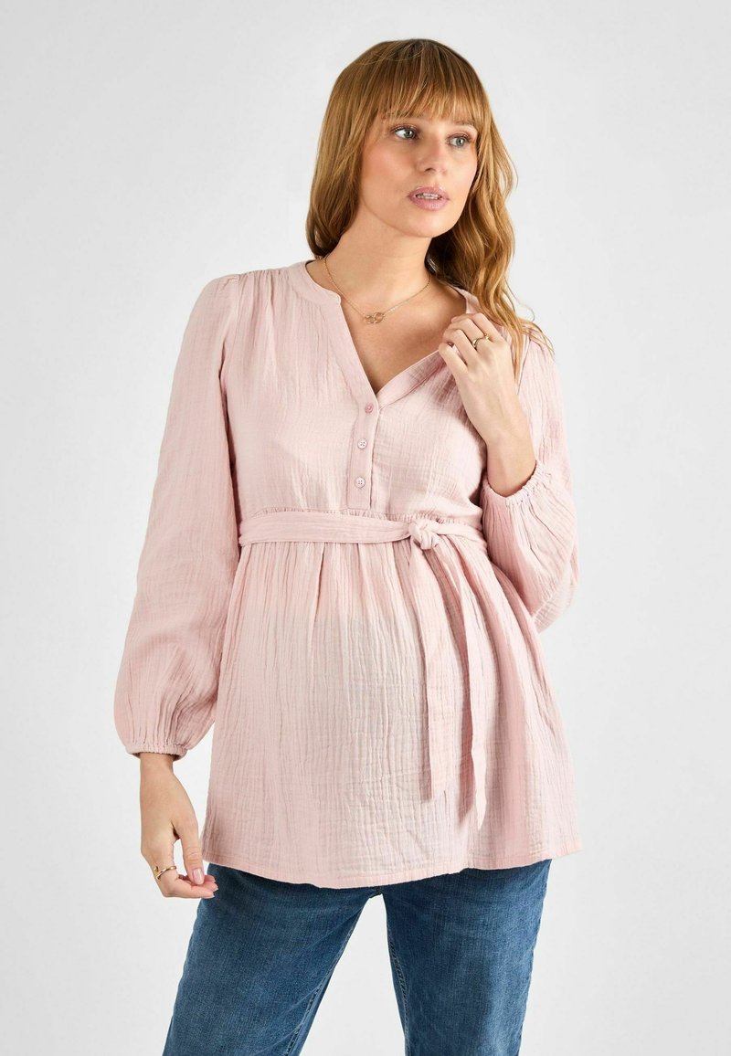 Blusa rosa chiaro realizzata in tessuto texturizzato, con scollo a V bottonato, maniche lunghe elasticizzate e una cintura annodata in vita.