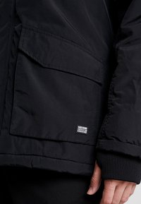 Mustapuku-tasku, jossa on läppä ja pieni Carsjeans Supply Goods -tunnus, henkilön käsi ja ribbikuvioitu hihansuu näkyvissä.