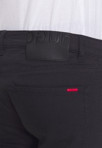 Pantalones de mezclilla negros con una cinturilla de cuero negro liso con logo en relieve y un pequeño acento de marca roja en el bolsillo trasero.