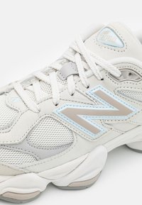 Valkoiset tennarit, joissa on verkko- ja nahkamateriaaleja, sininen ja hopea "N"-logo, rakenteelliset yksityiskohdat ja nauhat. Pyöreä kärki ja pehmustettu pohja.