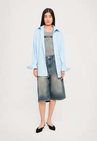 Mladá žena oblečená v svetlomodrej košeli s gombíkmi cez šedý top Moschino Jeans, širokých džínsových šortkách a čiernych sandále s podpätkami, stojaca na bielom pozadí.