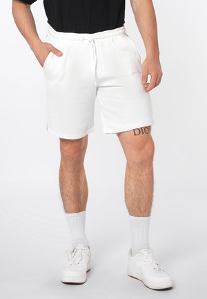 Mann trägt weiße Shorts mit Kordelzug, weiße Socken und weiße Turnschuhe, mit sichtbarem Tattoo am oberen linken Oberschenkel.