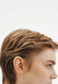 Cabello corto castaño con capas texturizadas, luciendo un pendiente de aro rectangular dorado, contra un fondo blanco liso.