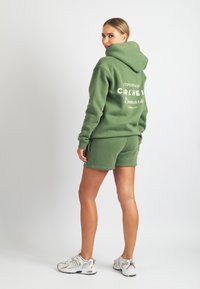 Zaļš sporta krekls ar kapuci un atbilstoši zaļas šortas no mīksta auduma, ar baltu tekstu, brīva piegriezuma. Valkāts ar gaišām sporta čībām.