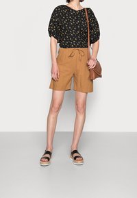 Blusa nera a fiori con maniche a sbuffo, pantaloni corti beige con coulisse e sandali neri con plataforma. Borsa marrone con dettaglio di frange.