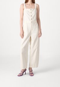 Krämfärgad jumpsuit med fyrkantig halsringning, vida ben, framknappar och justerbara axelband. Bärs med lila klackar med remmar.