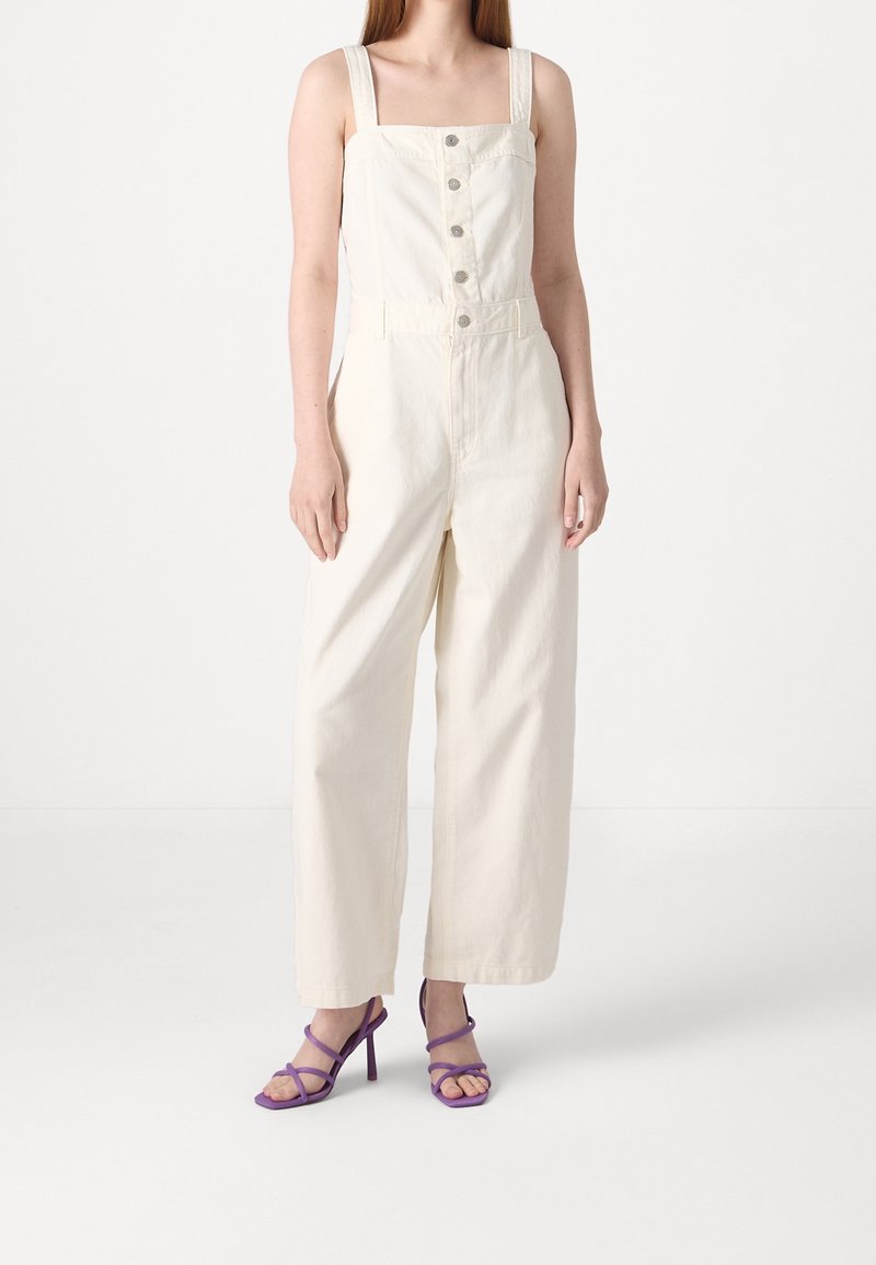 Krämfärgad jumpsuit med fyrkantig halsringning, vida ben, framknappar och justerbara axelband. Bärs med lila klackar med remmar.