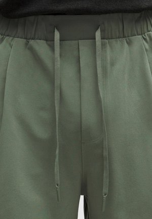 Oliivinvihreät vetoköydellä varustetut collegehousut, musta elastinen vyötärönauha ja pitkät, litteät narut roikkuvat edessä.