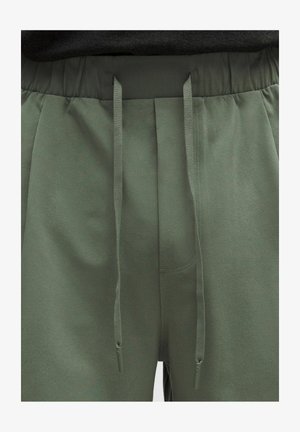 Oliivinvihreät vetoköydellä varustetut collegehousut, musta elastinen vyötärönauha ja pitkät, litteät narut roikkuvat edessä.