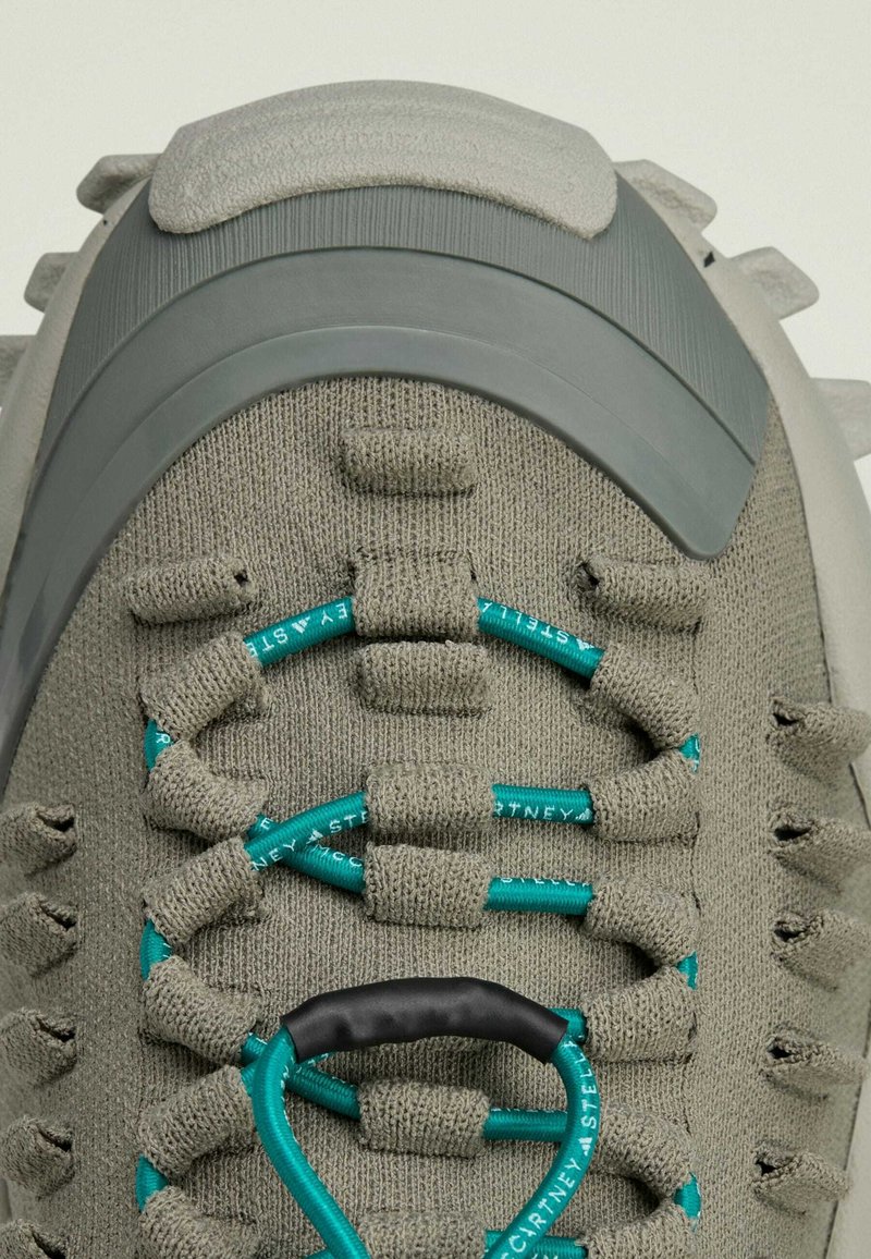 Grijze textielsneaker met een gestructureerd oppervlak, voorzien van turquoise veters en een gevormde rubberen hak. Opmerkelijk door het unieke vetersysteem.