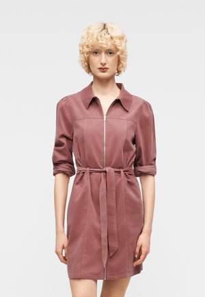 Persona rubia con vestido rosa polvoriento de manga remangada, cuello, cremallera frontal y cinturón atado a la cintura, de pie contra un fondo claro y liso.