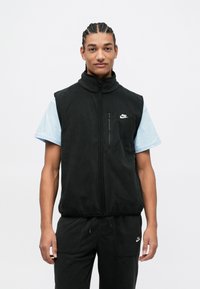 Vest de fleece negru cu guler înalt, fermoar frontal și buzunar pe pieptul stâng. Purpă peste o cămașă cu mânecă scurtă de un albastru deschis, asortată cu pantaloni închiși la culoare.