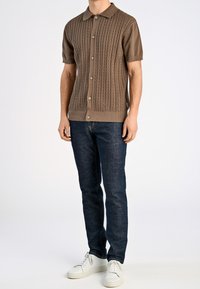 Mies, joka on pukeutunut ruskeaan lyhythihaiseen neulepaitaan, tummansinisiin farkkuihin ja valkoisiin lenkkareihin, seisoo valkoista taustaa vasten.