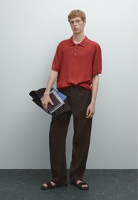 Červené polo tričko s krátkým rukávem a se třemi knoflíky, v kombinaci s volnými hnědými kalhotami. Model drží tmavou tašku a knihy, na nohou má černé sandály.