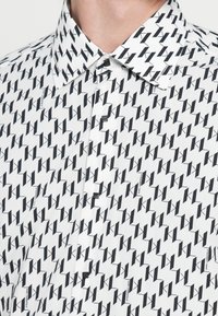 Fehér ing, amelyen merész, fekete, ismétlődő "K" mintázat látható. Klasszikus gallér, gombos lezárás, a szövet textúrája sima és könnyűnek tűnik.