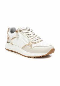 Zapatilla blanca y beige con una superficie texturizada, acentos metálicos, cierre con cremallera y suela de goma. Presenta perforaciones en la parte delantera.
