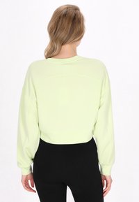 Felpa corta verde chiaro con maniche lunghe e oversize, caratterizzata da un colletto rotondo e una texture liscia. Indossata con leggings neri.