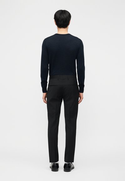 Man die zwarte pantalon, zwart shirt met lange mouwen, witte sokken en zwarte nette schoenen draagt, staand met de rug naar voren tegen een effen achtergrond.