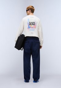 Κρεμ πουκάμισο μακρυμάνικο με πολύχρωμο γραφικό στην πλάτη, συνδυασμένο με μπλε σκούρο παντελόνι φαρδύ και μαύρη τσάντα.